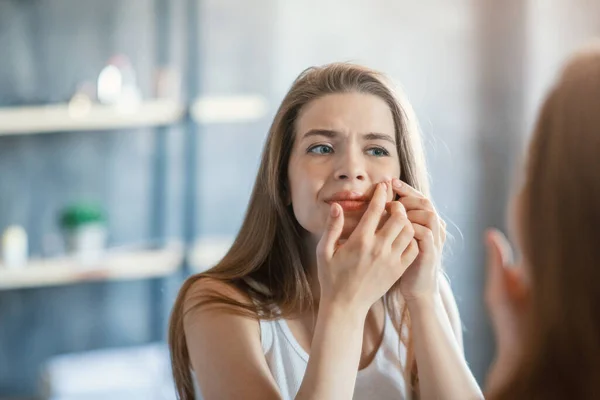 Unhappy millennial woman squeezing her acne near looking glass at home, empty space — Stock Photo, Image