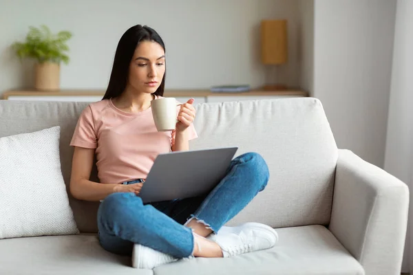 Ragazza utilizzando computer portatile avendo caffè seduto sul divano a casa — Foto Stock