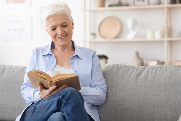 Kedvenc időtöltés. Mosolygó Idős hölgy Olvasmány Könyv Kényelmes kanapé Otthon — Stock Fotó