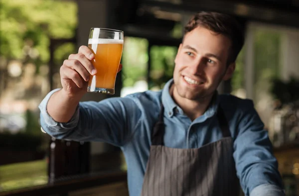 Färsk dryck av hög kvalitet. Nöjd leende bartender i förkläde höjer glas av ljus ale upp och ser på sin färg — Stockfoto