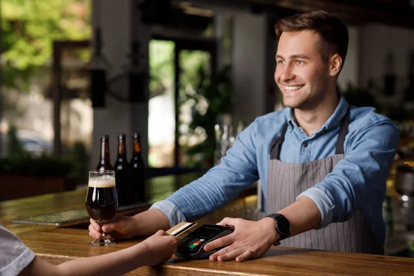 Barman com terminal e copo de cerveja escura, olha para o cliente com cartão de crédito — Fotografia de Stock