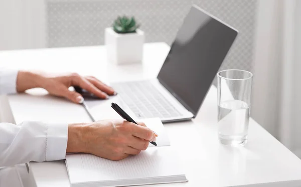 Female hands make entry in notebook on table with laptop with blank screen