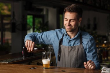 Müşteri için bira doldurur. Önlüklü gülümseyen barmen elinde bir şişe bira tutuyor.