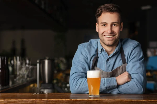 Tradições de cerveja no pub moderno. Sorrindo cara bonito na camisa jeans inclina-se no balcão de bar com vidro — Fotografia de Stock