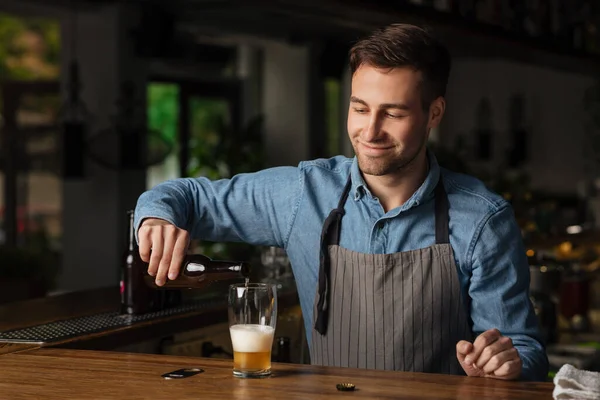 Schenkt bier in voor de klant. Lachende barman in schort houdt fles pils vast — Stockfoto