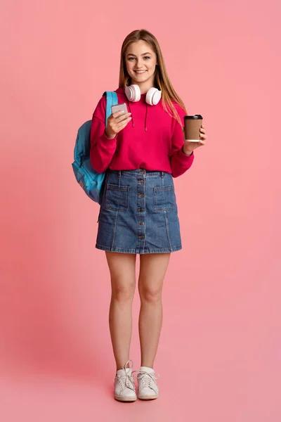 Сучасна дівчина-підліток з чашкою кави, рюкзаком та навушниками, перевірка на смартфон — стокове фото