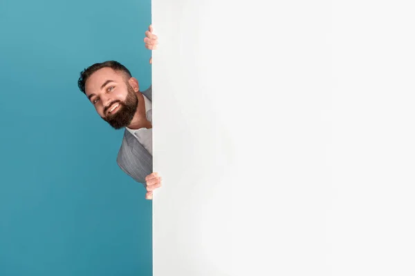 Empresario sonriente escondido detrás de un póster en blanco con espacio para texto o diseño sobre fondo azul —  Fotos de Stock
