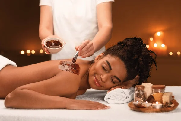 Terapeuta feminina aplicando máscara esfoliante no corpo da mulher africana — Fotografia de Stock