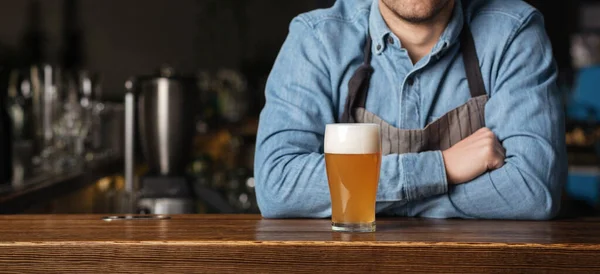 Cerveza. Cantinero con brazos cruzados, descansando sobre barra de madera, con vaso de cerveza ligera con espuma — Foto de Stock
