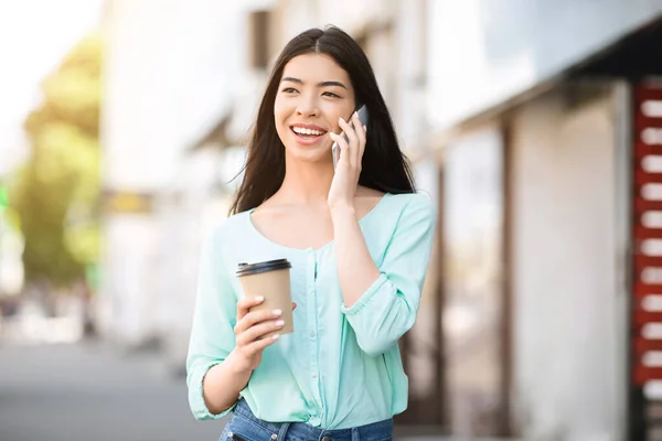 Comunicación moderna. Asiática chica caminando al aire libre con café y hablando en el teléfono celular — Foto de Stock