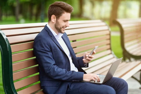 Pracownik biurowy z laptopem i telefonem komórkowym siedzący na ławce w parku i kończący projekt — Zdjęcie stockowe