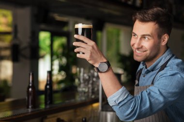 İyi iş çıkardın. Modern barmen gülümser ve koyu renkli bira ile yükseltilmiş bardağa bakar.