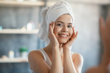 Pretty girl applying under eye cream on her perfect skin after bath near mirror indoors clipart