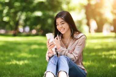 Şehir parkında akıllı telefondan müzik dinleyen mutlu Asyalı kadın.