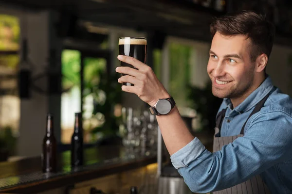 Goed gedaan. Moderne barman glimlacht en kijkt naar hoog verhoogd glas met donker ambachtelijk bier — Stockfoto