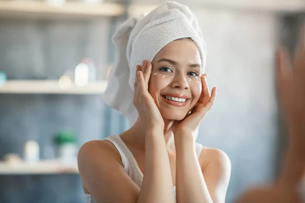 Chica bonita aplicando bajo la crema para los ojos en su piel perfecta después del baño cerca del espejo en el interior — Foto de Stock