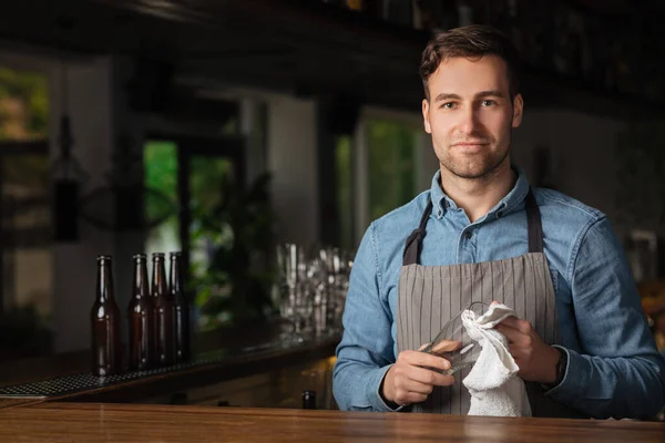 Modern werk van barman. Knappe man in schort met stijlvol kapsel, rubs glas — Stockfoto