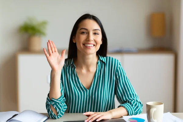 Счастливая деловая женщина машет привет улыбаясь на камеру сидя в офисе — стоковое фото