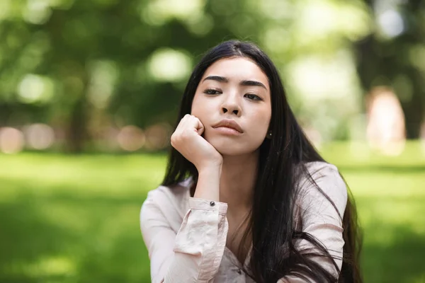 Pensativa asiática chica sentado en el césped en parque y mirando lejos, primer plano —  Fotos de Stock