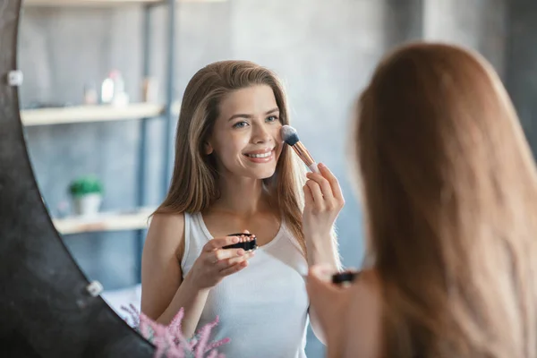 Όμορφη νεαρή γυναίκα εφαρμογή blusher με πινέλο καλλυντικών μπροστά από τον καθρέφτη στο μπάνιο — Φωτογραφία Αρχείου