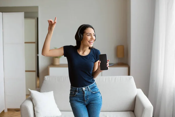 女性の歌と踊り自宅でスマートフォンを保持するヘッドフォンを身に着けている — ストック写真