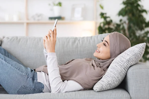 Relajada chica árabe en Hijab Descansando en el sofá con el teléfono inteligente en las manos — Foto de Stock