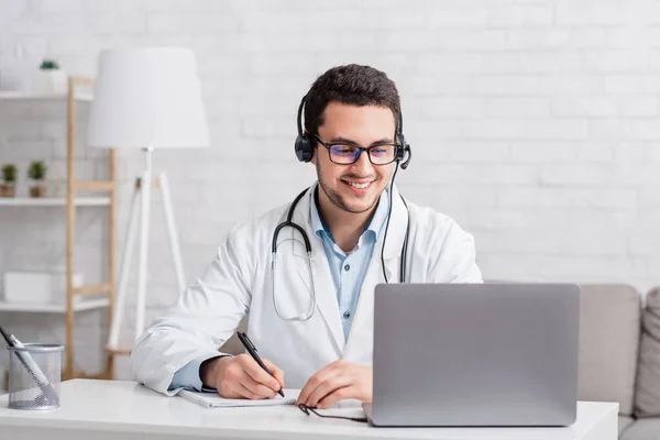 Glimlachende arts in witte jas met hoofdtelefoon, maakt notities in notebook kijkt in laptop — Stockfoto