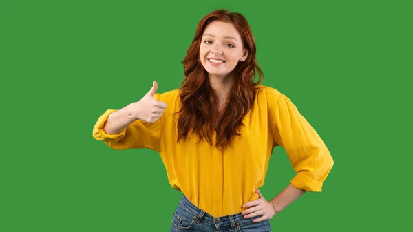 Červenovlasá dívka gestikulující palce nahoru s úsměvem na fotoaparát stojící ve studiu — Stock fotografie