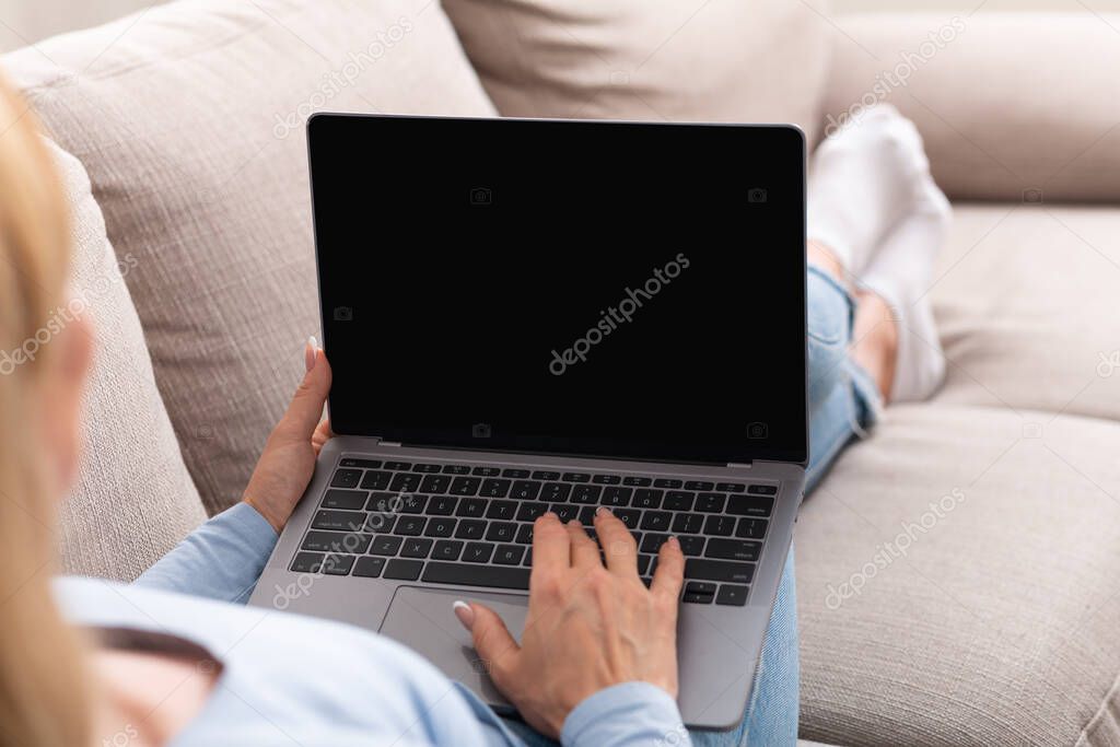 Digital technology for work from home. Woman typing on laptop with blank screen, lying on sofa in home interior, shoulder view
