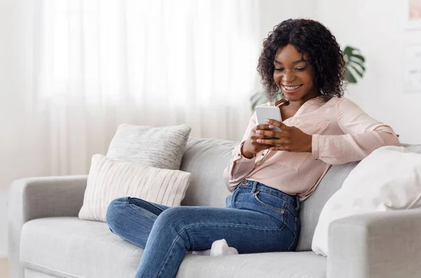 Sorrindo mensagens menina preta no smartphone em casa, relaxando no sofá confortável — Fotografia de Stock