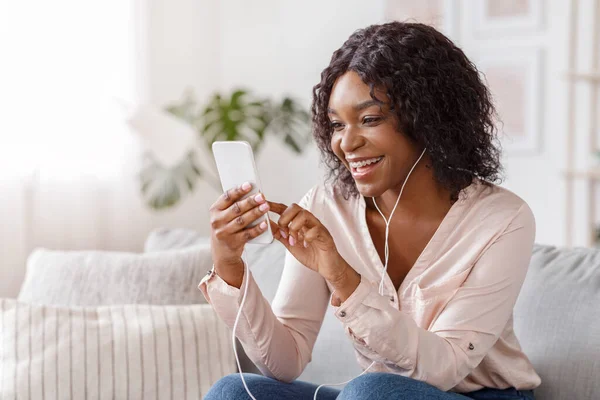Mensagens menina preta feliz no smartphone e ouvir música em casa — Fotografia de Stock