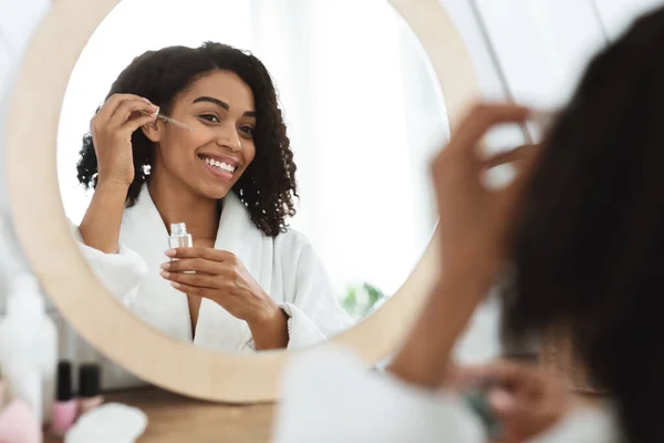 Улыбающаяся черная женщина наносит увлажняющую сыворотку лицом к лицу и смотрит в зеркало — стоковое фото