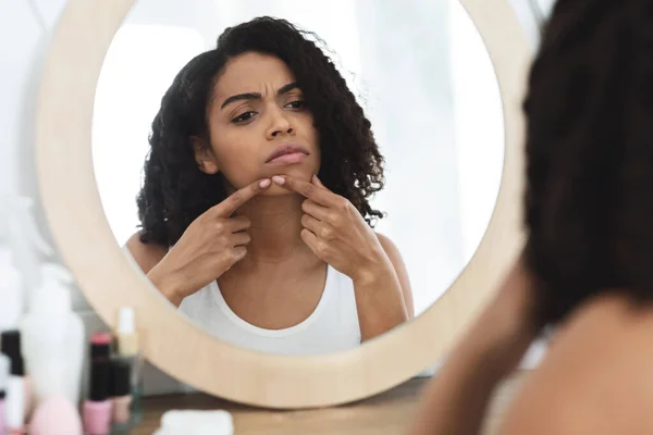 Концепция проблемной кожи. Афроамериканская девушка смотрит в зеркало и трогает прыщ — стоковое фото