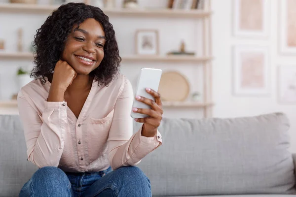 App fixe. Mulher negra bonita sentada no sofá e usando smartphone — Fotografia de Stock