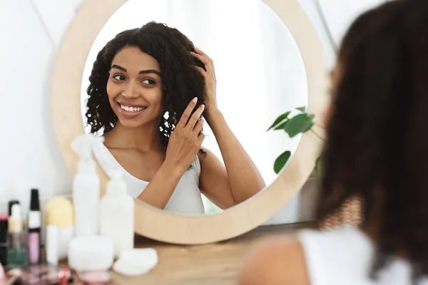 Belleza natural. Feliz chica negra tocando su suave pelo rizado cerca del espejo — Foto de Stock