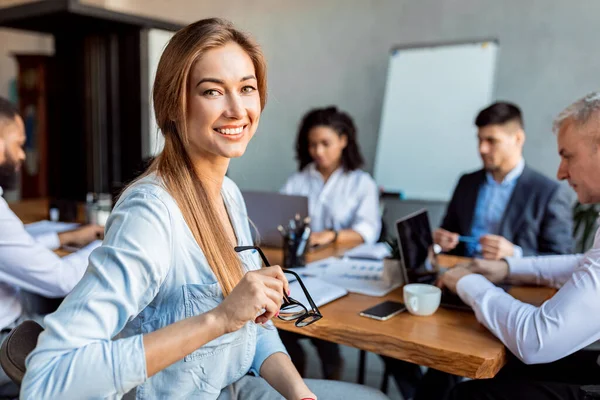 Happy Businesswoman při pohledu na kameru sedí s kolegy v kanceláři — Stock fotografie