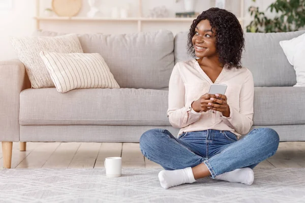 Dromerig zwart meisje ontspannen op de vloer thuis met smartphone en koffie — Stockfoto