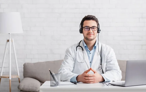 Personlig blogg av läkare. Man med hörlurar, sitter vid bord med laptop i interiören — Stockfoto