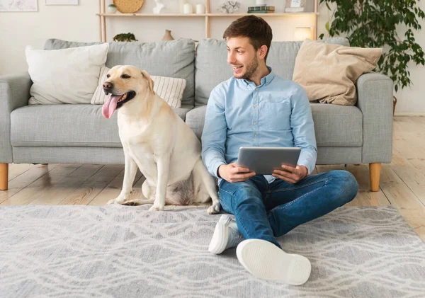 Evde elinde tablet ve labrador olan genç bir adam var.