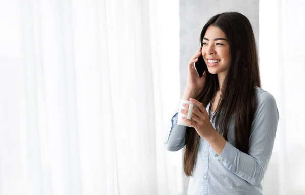 Hoş bir karar. Cep telefonuyla konuşan ve pencerenin yanında kahve içen Asyalı kız. — Stok fotoğraf