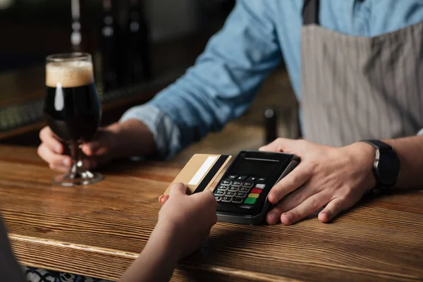 Negócios de bar. Barman no avental dá vidro de cerveja e terminal, menina paga conta com cartão — Fotografia de Stock