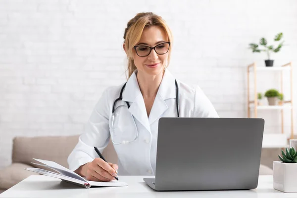 Пациент разговаривает с доктором по видеосвязи на ноутбуке. Женщина в белом халате делает заметки в блокноте — стоковое фото