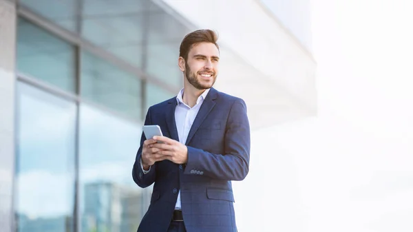 Sonriente industrial milenario revisando su teléfono inteligente cerca de rascacielos en la calle de la ciudad — Foto de Stock