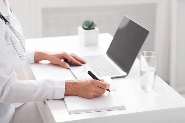 Online-Kommunikation mit Patienten und Notizen. Ärztin schreibt in Notizbuch am Tisch mit Laptop — Stockfoto