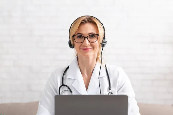 Linha directa no hospital. Médico sorridente em casaco branco, em fone de ouvido com laptop — Fotografia de Stock