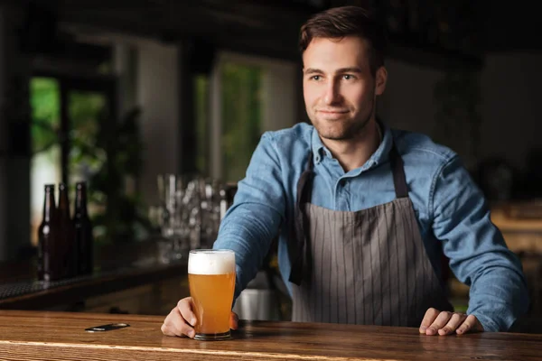 Fiesta de cerveza artesanal. Barman guapo en delantal detrás de la barra con un vaso de cerveza ligera — Foto de Stock