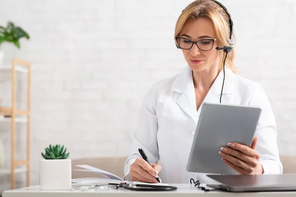 Рабочее место онлайн медицинского консультанта. Женщина с планшетом в руках делает заметки в блокноте — стоковое фото