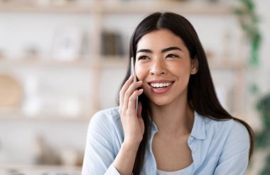 Modern İletişim. Neşeli Asyalı kız evde cep telefonuyla konuşuyor, sohbetten zevk alıyor.
