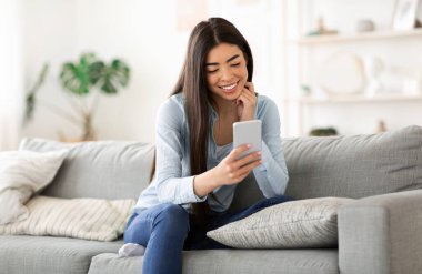 Genç Asyalı kız akıllı telefonuyla mesajlaşırken evdeki kanepede dinleniyor.