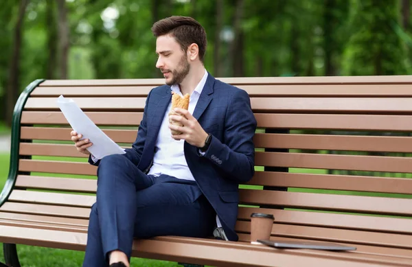 Hardworking CEO looking through documents while having lunch on bench at park — Stock Photo, Image